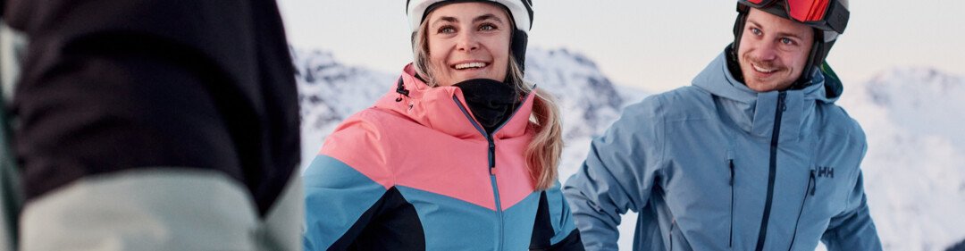 Three skiers wearing helmets and winter jackets on a snowy mountain slope, a woman in a pink and blue jacket smiles in the foreground, a man in a blue jacket stands to the right and looks at her. | © INTERSPORT International Corporation GmbH