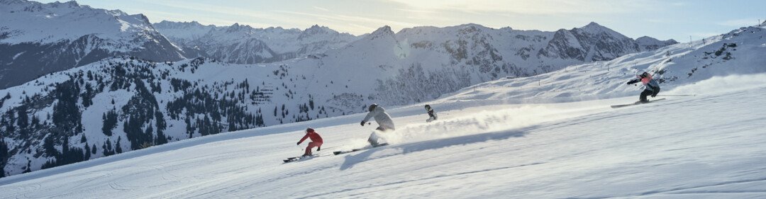 Four skiers in winter sports clothing ski down a sunny, freshly groomed slope in single file; snow-covered Alpine mountains in the background. | © INTERSPORT International Corporation GmbH