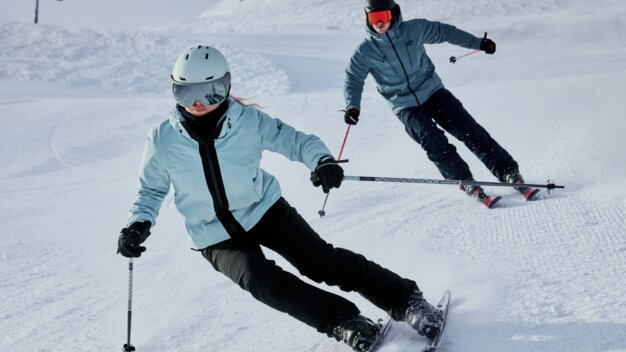 Two skiers in light blue jackets are skiing energetically down a groomed ski slope; in front is a female skier with a white helmet and mirrored ski goggles, followed by a male skier with a black helmet. | © INTERSPORT International Corporation GmbH