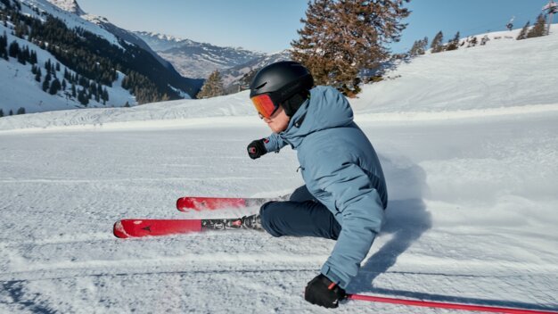 Skiers in blue anoraks and black helmets with red skis carve turns on a groomed slope in front of snow-covered mountains. | © INTERSPORT International Corporation GmbH