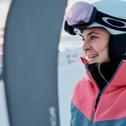 Frau in weißem Skihelm und Skibrille, lächelnd in pink-blauer Skijacke neben einem Ski vor verschneiter Berglandschaft. | © INTERSPORT International Corporation GmbH