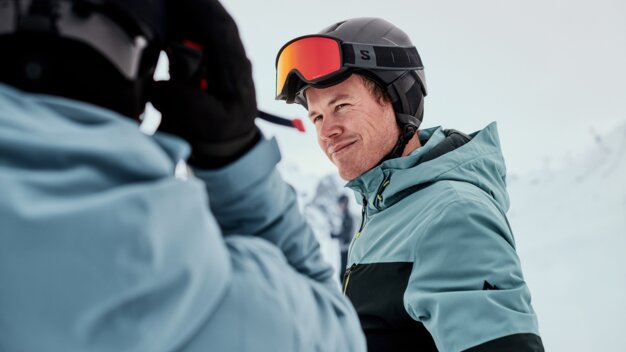 Two people skiing: close-up of a smiling man in a blue-green ski jacket with a helmet and orange ski goggles, the shoulder of a second person in the foreground, snow-covered mountain landscape in the background. | © INTERSPORT International Corporation GmbH