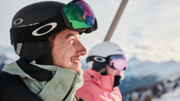 Two skiers on a chairlift: close-up of a smiling man wearing a black helmet and mirrored ski goggles, with a woman wearing a white helmet and pink ski jacket in the background, set against snow-covered mountains. | © INTERSPORT International Corporation GmbH