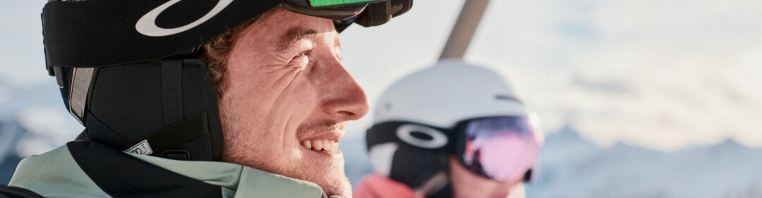 Two skiers on a chairlift: close-up of a smiling man wearing a black helmet and mirrored ski goggles, with a woman wearing a white helmet and pink ski jacket in the background, set against snow-covered mountains. | © INTERSPORT International Corporation GmbH