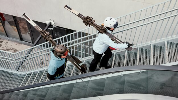 Two people wearing helmets and winter jackets carry skis over their shoulders and climb a metal outdoor staircase, view from above, urban environment. | © INTERSPORT International Corporation GmbH