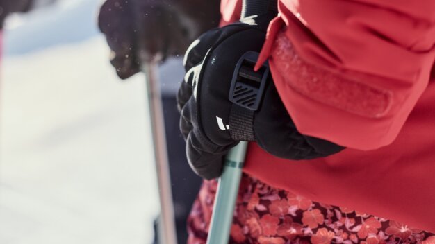 Close-up of a black-gloved hand holding a ski pole; red ski jacket and floral ski pants, snowy mountain landscape in the background. | © INTERSPORT International Corporation GmbH