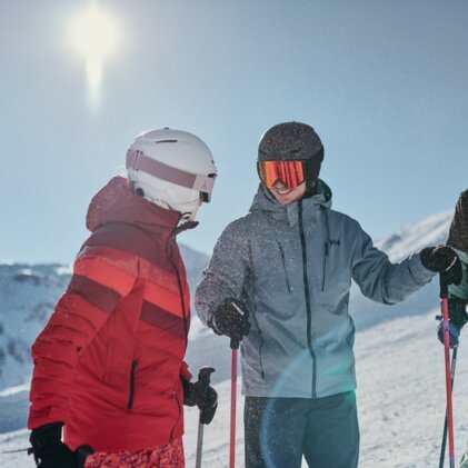 Three skiers in winter jackets and helmets stand on a snowy slope, laughing and leaning on ski poles; sunny mountain backdrop in the background. | © INTERSPORT International Corporation GmbH