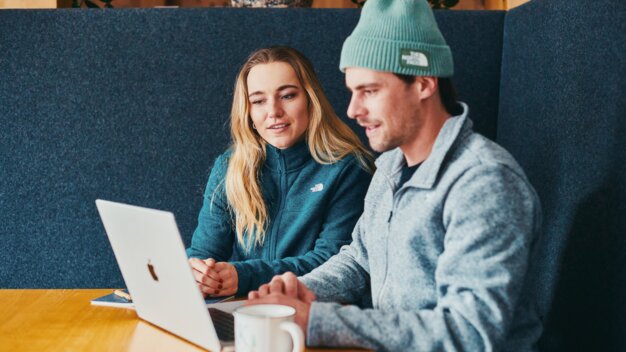 Zwei Personen in einer dunklen Sitznische sitzen an einem Holztisch und schauen auf einen offenen Laptop; beide tragen warme Fleecejacken (The North Face) und eine Kaffeetasse steht auf dem Tisch. | © INTERSPORT International Corporation GmbH