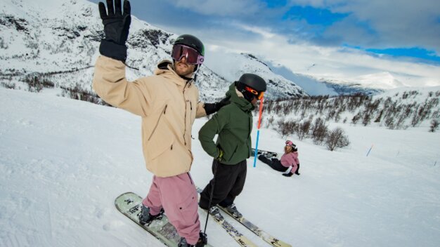 Ein Mann auf einem Snowboard steht auf der Piste neben einem Skifahrer und hebt die Hand zum Winken.
