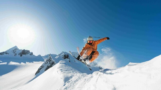 Skiers in orange gear with helmets and mirrored ski goggles jump over a snowy ridge in front of snow-covered mountains and blue skies. | © INTERSPORT International Corporation GmbH
