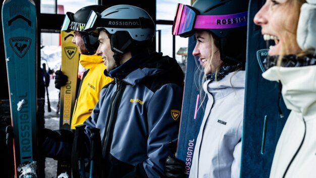 Four people wearing ski helmets and winter jackets with the Rossignol logo, standing side by side and smiling, holding their skis vertically; winter mountain scenery in the background. | © INTERSPORT International Corporation GmbH