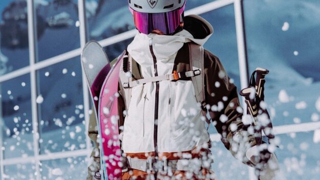 Skier wearing a white jacket, helmet, and purple ski goggles carries pink skis and poles; snow sprays up in front of a glass facade with mountain reflections. | © INTERSPORT International Corporation GmbH