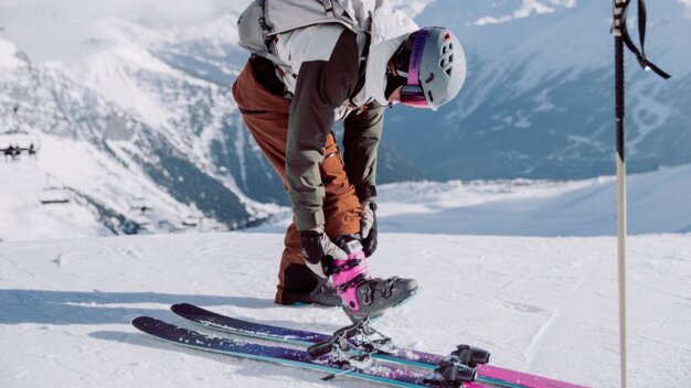 Person wearing a ski helmet and backpack, attaching a pink and black ski boot buckle to a binding on a snow-covered mountain slope; looking down, mountains in the background. | © INTERSPORT International Corporation GmbH