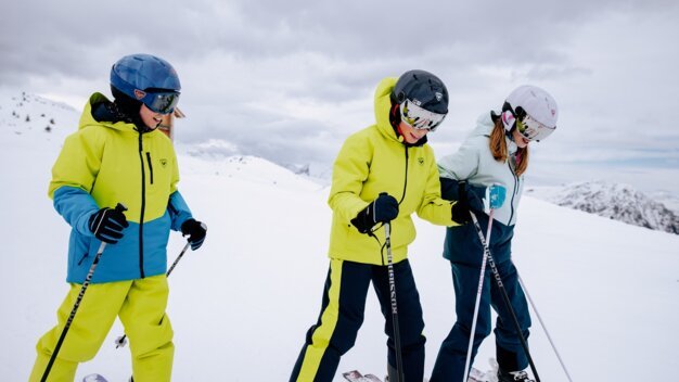 Three children wearing helmets and colorful ski jackets stand laughing side by side on skis on a snowy mountain slope. | © INTERSPORT International Corporation GmbH