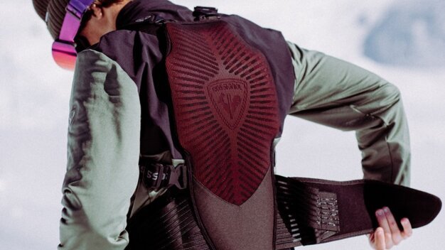 Person seen from behind putting on a padded back protector/airbag vest over orange ski pants; wearing a green jacket, brown hat, and purple ski goggles in front of a snowy mountain landscape. | © INTERSPORT International Corporation GmbH