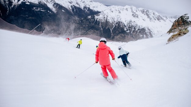 Three skiers on a snowy slope, one person in the foreground wearing a red ski jacket and helmet with ski poles, snow-covered mountains and safety net in the background | © INTERSPORT International Corporation GmbH