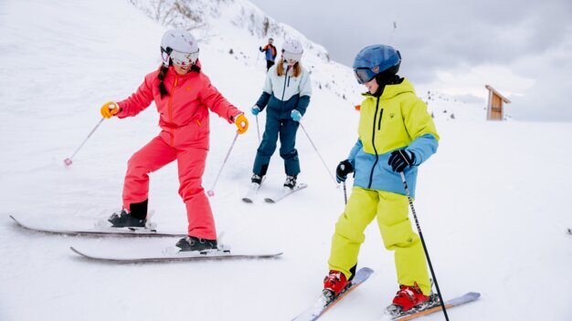 Three children in colorful ski suits (red, yellow, blue) with helmets and ski poles skiing on a snowy slope against a mountainous backdrop. | © INTERSPORT International Corporation GmbH