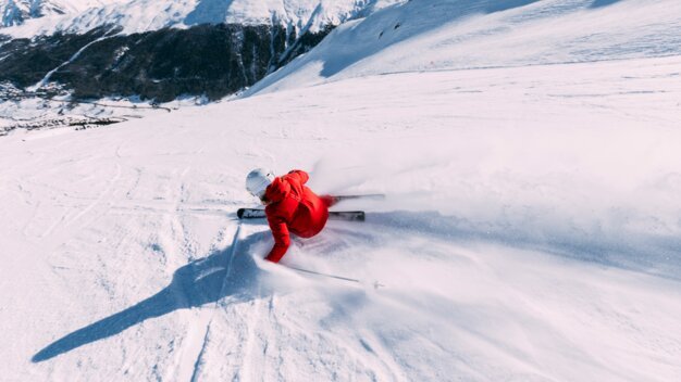 Skiers in red jackets and white helmets carving down the slope, powder snow spraying, snow-covered mountain backdrop in the background. | © INTERSPORT International Corporation GmbH