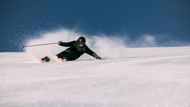 Skiers in black gear and helmets with orange ski tips carving, snow spray on groomed slopes against a clear blue sky. | © INTERSPORT International Corporation GmbH