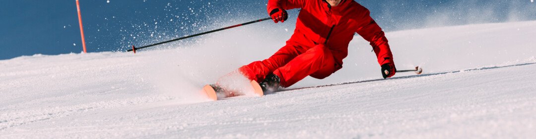 Skiers in red jackets and helmets dynamically descend a groomed slope, snow spraying up, blue sky in the background. | © INTERSPORT International Corporation GmbH