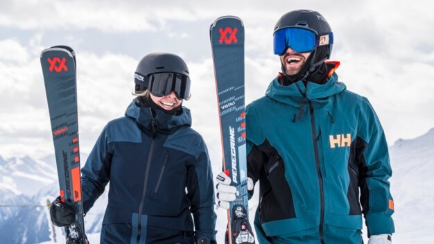 Two smiling skiers wearing helmets and ski goggles are each holding a Völkl ski in front of a snowy mountain landscape. | © INTERSPORT International Corporation GmbH