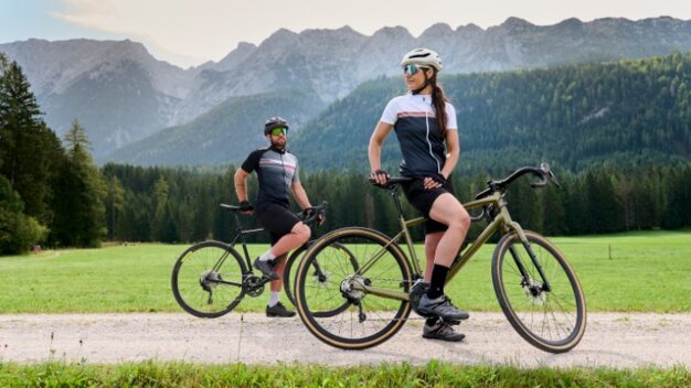 Zwei Radfahrer auf einem Schotterweg, umgeben von grünen Wiesen und einer eindrucksvollen Berglandschaft.