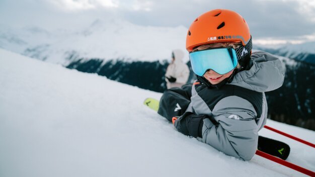 Kind mit orangefarbenem Skihelm und blauer Skibrille sitzt im Schnee.