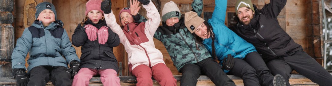 Gruppo di persone in abbigliamento invernale sedute di fronte a una baita di legno che salutano. | © INTERSPORT International Corporation GmbH