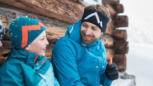 Vater und Sohn lächeln sich in Winterkleidung vor einer Holzhütte an. | © IIC/McKINLEY