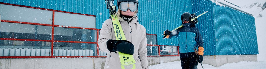 © Fischer Sports GmbH Due sciatori con gli sci davanti a un edificio blu nella neve. | © Fischer Sports GmbH