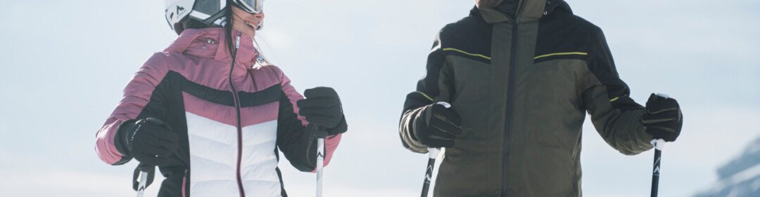 Due sciatori in abbigliamento da sci in piedi e sorridenti in una giornata nevosa. | © INTERSPORT International Corporation GmbH