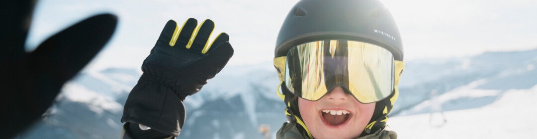 Ragazzo con maschera da sci e casco che saluta su una pista da sci. | © INTERSPORT International Corporation GmbH